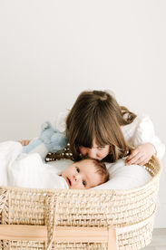 Newborn session in Guelph, Ontario featuring big sister kissing baby brother in a woven bassinet during a soft studio portrait