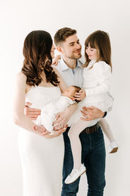Family of four during newborn session in Guelph, Ontario with mother holding swaddled newborn baby and father with young daughter in a bright studio setup