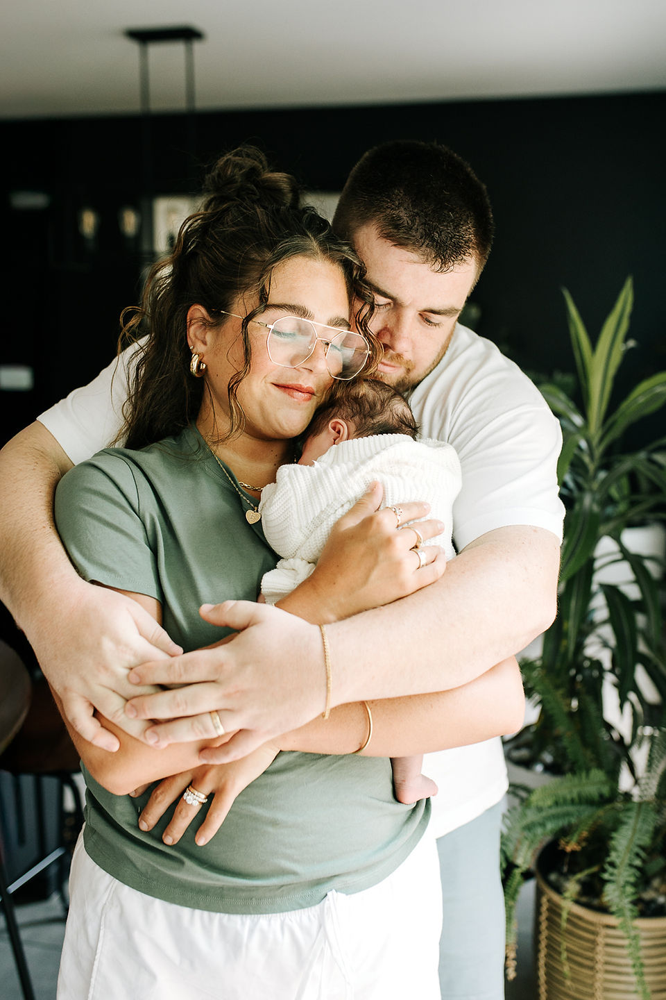 In-home newborn session in Guelph, Ontario featuring parents embracing while holding their newborn baby wrapped in a white blanket