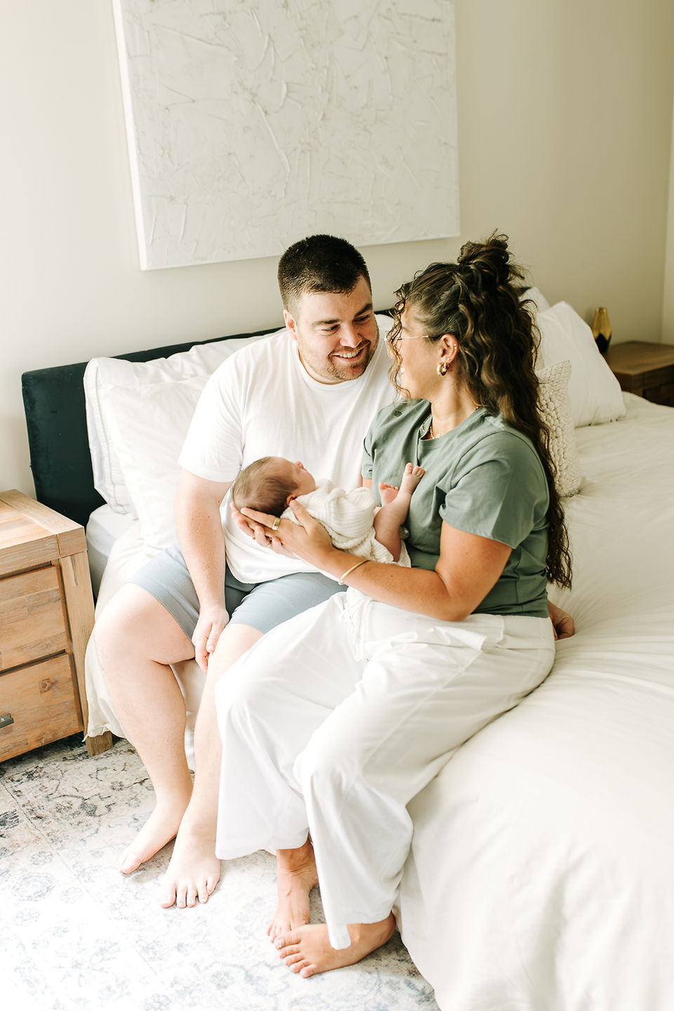 In-home newborn session in Guelph, Ontario featuring parents smiling at each other while cradling their newborn baby on the bed