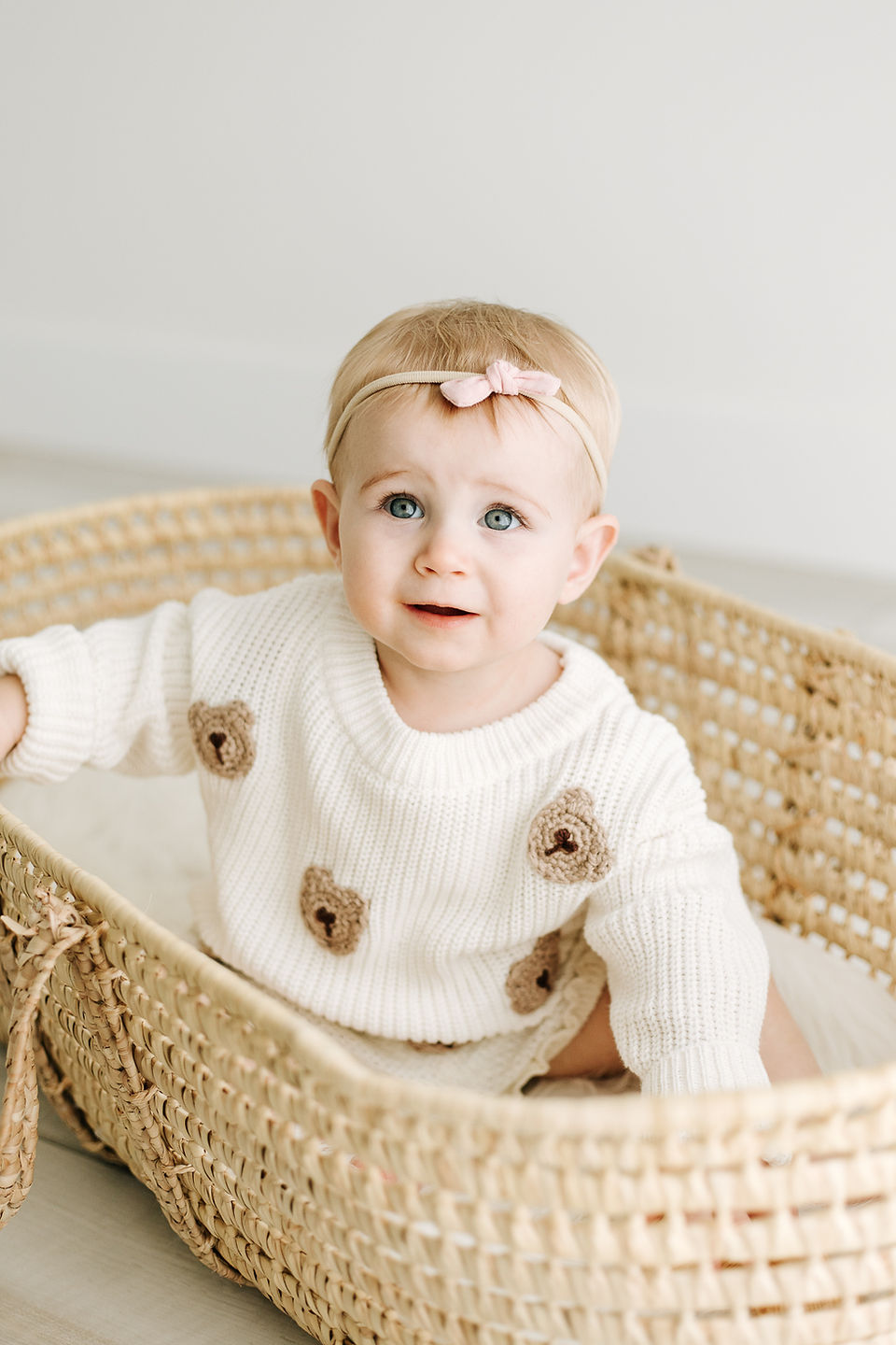 Studio milestone photography in Guelph, Ontario featuring a baby girl in a teddy bear sweater and pink bow headband sitting in a woven Moses basket.