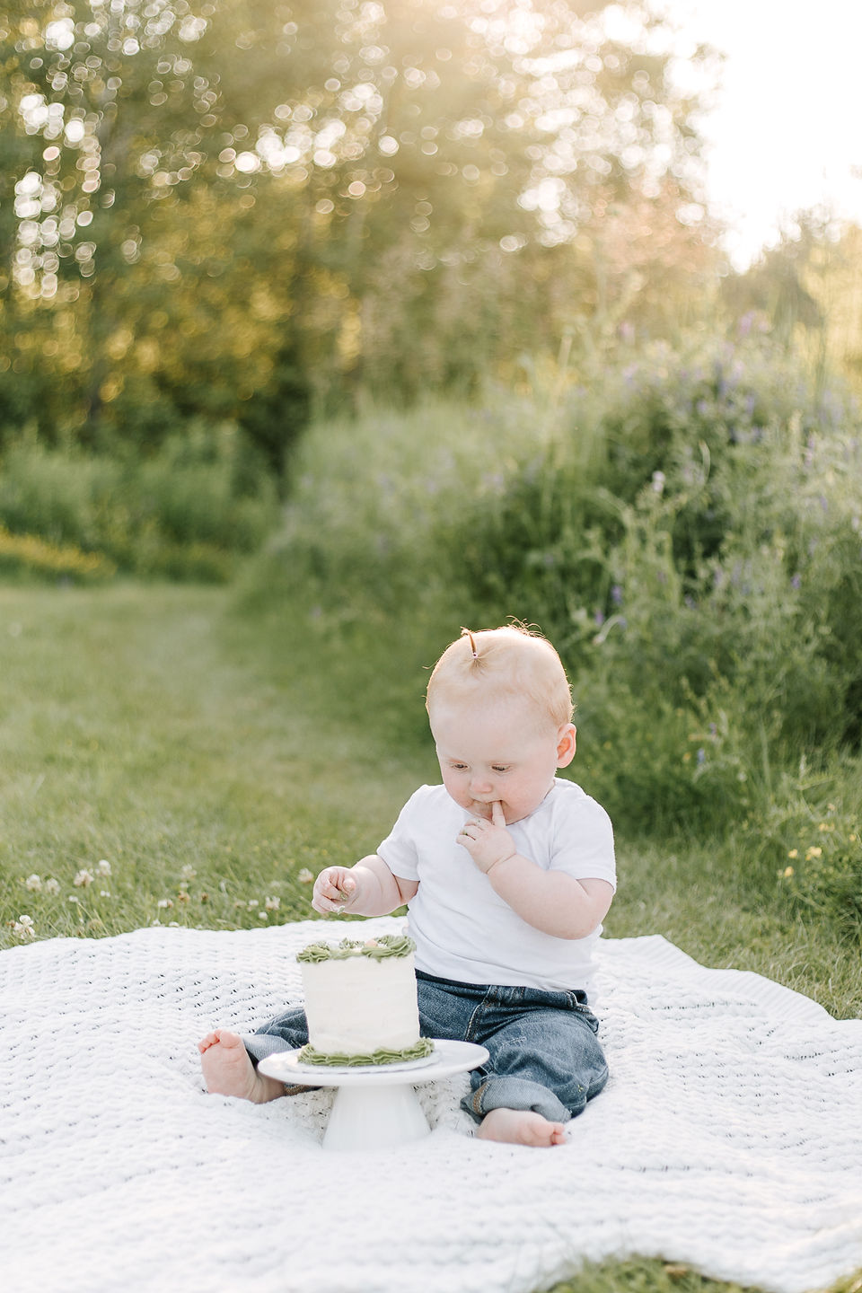 Baby tasting the frosting from her first birthday smash cake outdoors in Guelph, Ontario, wearing a white t-shirt and cuffed blue jeans while sitting on a white blanket in the grass during golden hour.