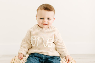 First birthday milestone photos of a baby boy in a ‘one’ sweater riding a toy race car at a studio s