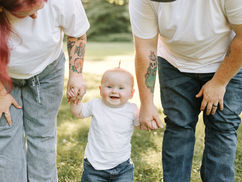 Young baby smiling between parents during an outdoor first birthday family session, wearing a white shirt and blue jeans while holding both parents’ hands on a sunny grassy lawn.