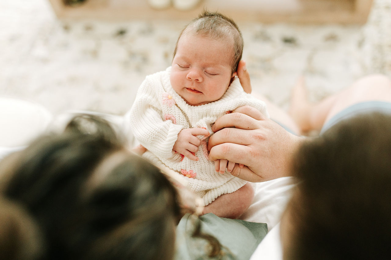 Newborn photography in Guelph, Ontario capturing a sleeping baby in a white sweater with pink floral details surrounded by mom and dad's loving hands
