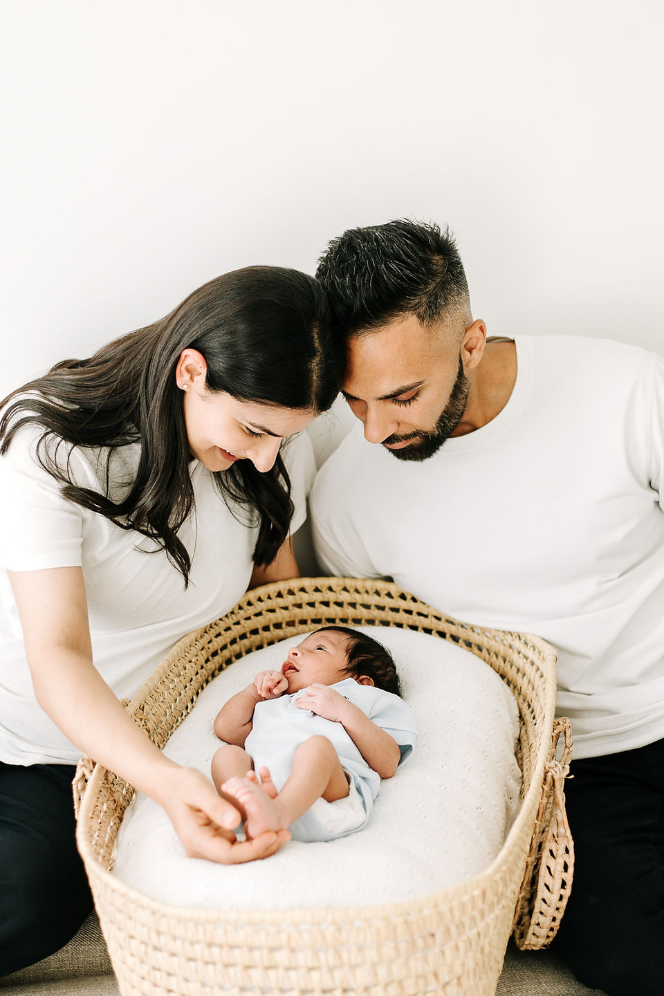 Studio newborn family portrait in Guelph, Ontario featuring parents in white shirts and black pants sitting beside their baby in a woven Moses basket