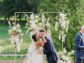 Bride and groom kissing under a floral arch at an outdoor wedding venue.