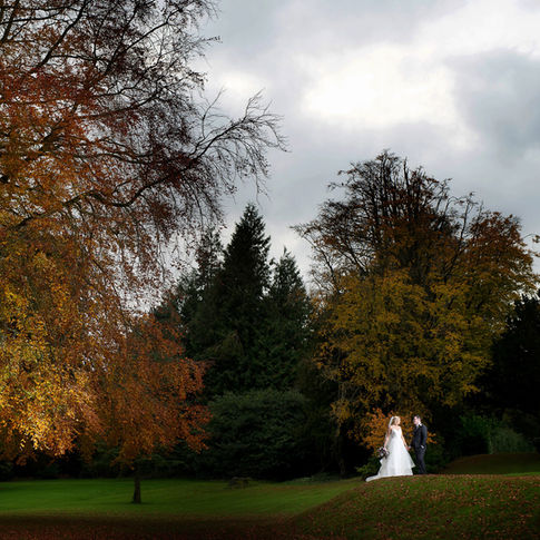 A bride and groom stand facing each other on a grassy hill surrounded by trees with autumn foliage at Bredenbury Court Barns. The sky is overcast, adding a moody atmosphere to the scene. Captured by Russell Lewis Photography, the bride is in a white gown, and the groom is in a dark suit.