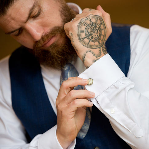 A bearded groom wearing a blue vest and a tie adjusts his cufflinks. His hand, adorned with a large clock tattoo, is positioned close to his shirt cuff. Captured by Herefordshire wedding photographer Russell Lewis Photography, the image highlights the intricate details of the tattoo and the act of fastening the cufflink.