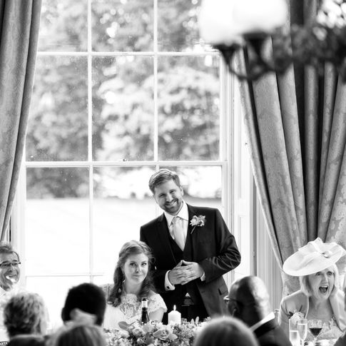 A black-and-white photo capturing a wedding reception at Homme House. The groom is standing and smiling while the seated bride looks content. Guests around them are engaged and happy. Outside the large window behind them, trees and a lawn are visible, beautifully shot by Herefordshire Wedding Photographer Russell Lewis