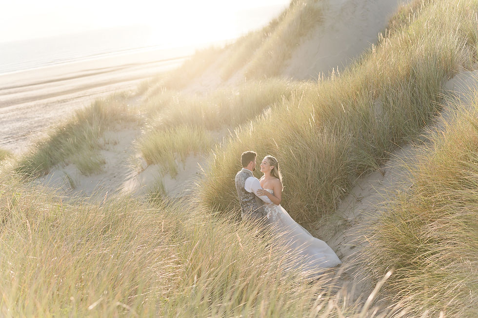 A couple stands embracing in a sunlit sandy dune with tall grass, overlooking a Aberdyfi beach. Romantic and tranquil atmosphere.