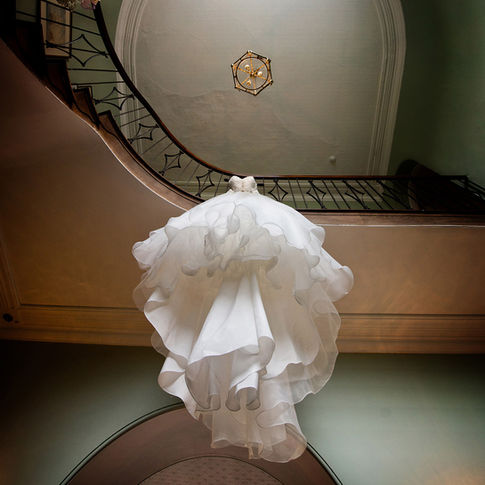 A white wedding dress with ruffled layers is suspended from a staircase railing, viewed from below. The dress is elegantly draped under a curved staircase at Homme House, captured by Herefordshire Wedding Photographer Russell Lewis. A gold hexagonal light fixture hangs from the ceiling.