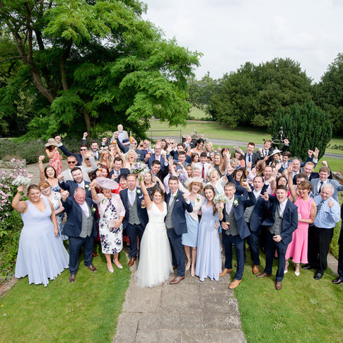 A large group of people, all dressed in formal attire, gather outside on a lush green lawn at Burton Court, celebrating a wedding. The bride and groom stand at the center of the group, smiling broadly, while everyone raises their hands and cheers in joy. Herefordshire Wedding Photographer Russell Lewis captures the moment perfectly. Trees and a pathway are in the background