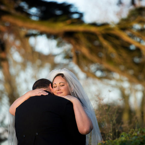 A bride and groom share an intimate embrace in an outdoor setting captured by Herefordshire Wedding Photographer Russell Lewis. The bride, wearing a white veil and dress, closes her eyes and smiles gently. The groom, in a black suit, faces away from the camera. The background features the natural light of Glewstone Court's gardens
