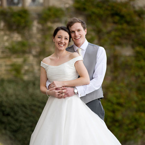 A couple poses together, smiling on their wedding day at Homme House. The bride wears an off-shoulder white wedding dress, while the groom is dressed in a light gray vest over a white shirt and gray pants. They stand in front of a greenery-covered stone wall, captured beautifully by Herefordshire Wedding Photographer Russell Lewis