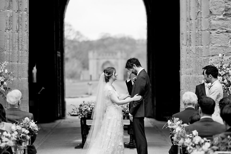 Bride and groom share an emotional moment in a stone archway, surrounded by guests and flowers. The groom wipes tears, creating a heartfelt scene at Hampton Court Castle in Herefordshire