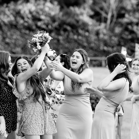 A black and white photograph captures a jubilant scene where a woman triumphantly raises a bouquet in the air, while others around her cheer and laugh. They are celebrating outdoors at Bredenbury Court Barns, with a lush background of trees and shrubs