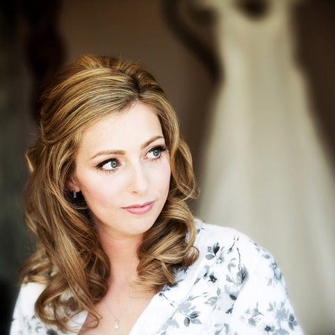 A bride with wavy blonde hair and blue eyes gazes off camera, wearing a patterned robe. Behind her, a white wedding dress hangs out of focus. The soft lighting highlights her elegant hairstyle and subtle makeup, giving the scene a serene and contemplative mood captured perfectly by Herefordshire Wedding Photographer Russell Lewis at Homme House