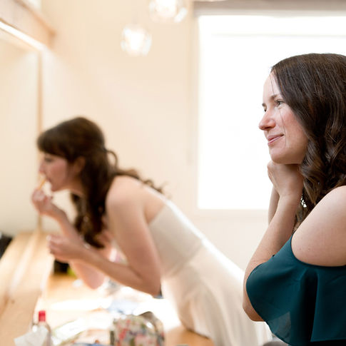 A bride and her bridesmaid get ready in the bridal room at Bredenbury Court Barns. Captured beautifully by Russell Lewis Photography, the bridesmaid is smiling with her hands near her face. She has long, wavy hair and is wearing a green off-shoulder dress. In the background, the bride, dressed in white, is applying lipstick while looking in a mirror