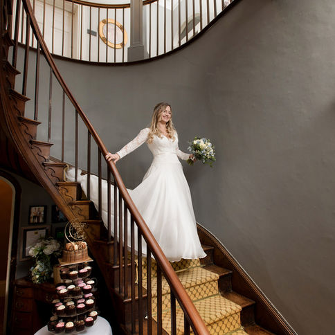 A bride, wearing a white gown with lace sleeves, smiles as she descends a curved wooden staircase at Glewstone Court. She holds a bouquet of flowers in one hand. Below the staircase, a table is set with an arrangement of cupcakes. Captured by Herefordshire wedding photographer Russell Lewis, the scene is elegant and warmly lit