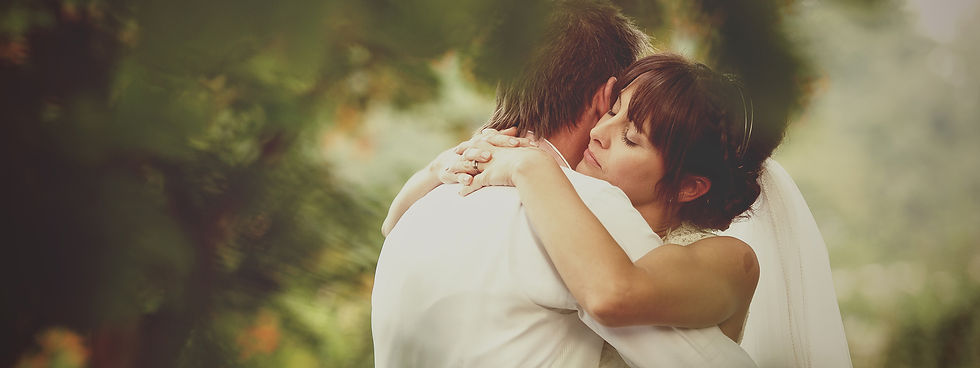 Bride and groom embrace tightly outdoors, eyes closed, in a soft-focus green garden, warm and romantic mood