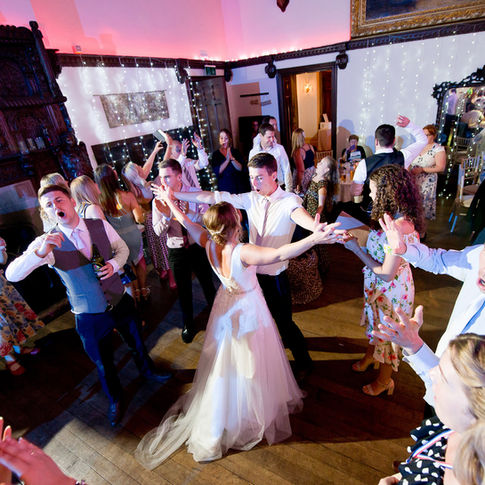 A bride and groom dance surrounded by guests at a Burton Court wedding reception. The room, captured beautifully by Herefordshire Wedding Photographer Russell Lewis, is decorated with string lights and ornate paneling. People are dancing energetically, singing along, and having a good time