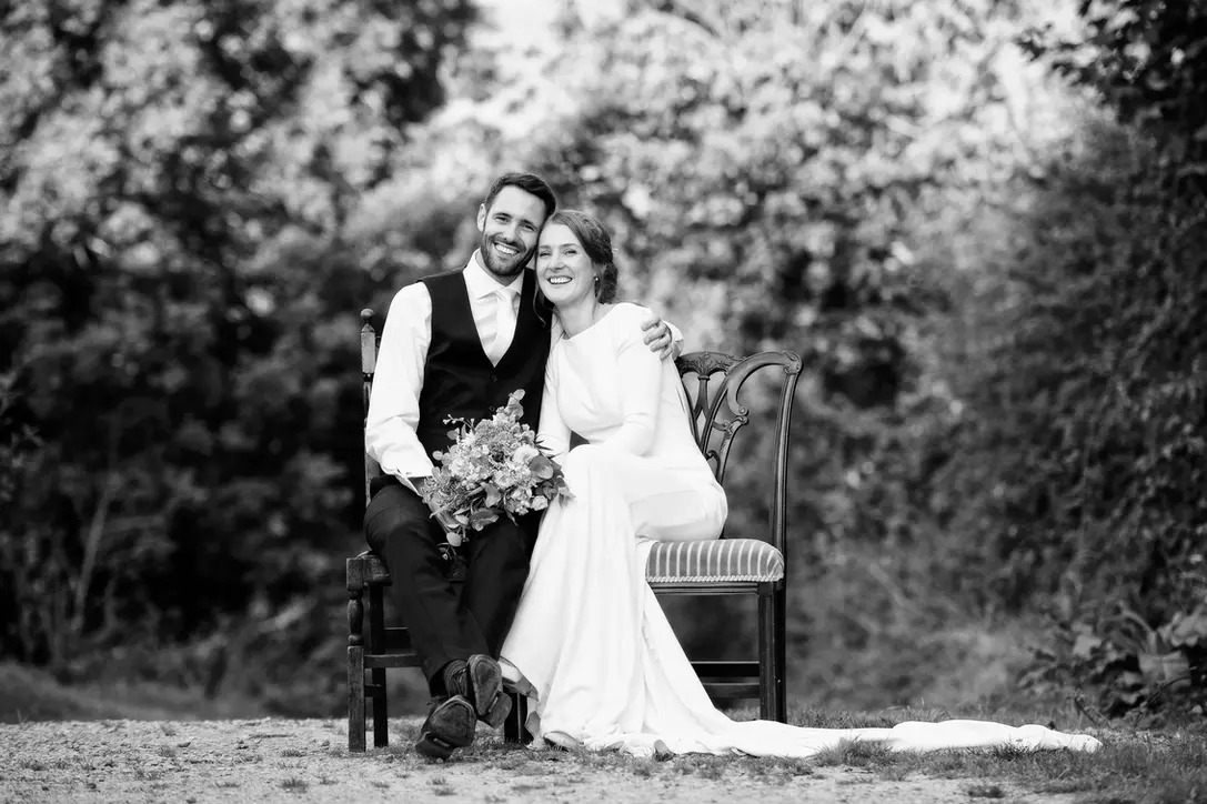 Bride and groom sitting together on wooden chairs outdoors during wedding portraits, photographed in black and white at Lyde Court in Herefordshire