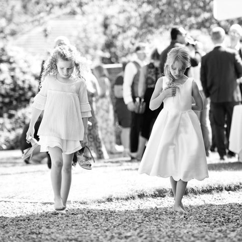 Black-and-white photo of two young girls walking barefoot on pebbled ground at Glewstone Court. The girl on the left has curly hair and is wearing a light-colored dress. The girl on the right has straight hair and is in a sleeveless dress, holding something close to her chest. People are in the background. Captured by Herefordshire wedding photographer Russell Lewis