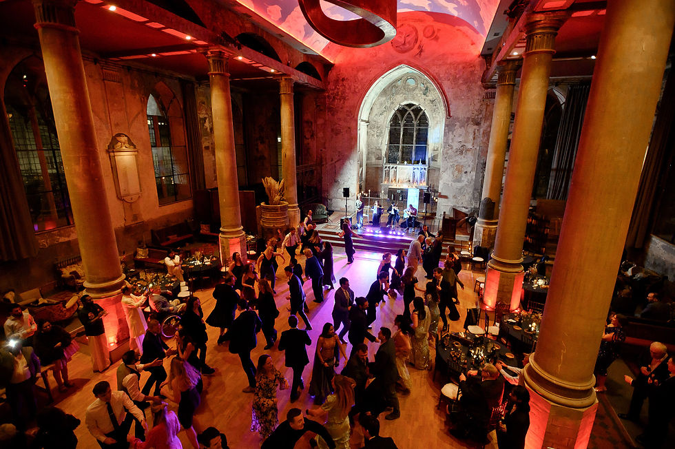 Elegant ballroom with dancing guests under colorful lighting. Band playing on stage; large columns and high ceiling create a grand atmosphere.