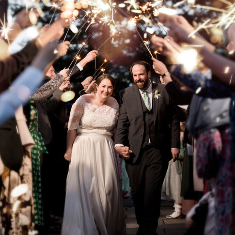 A newlywed couple, smiling and holding hands, walk through an archway of sparklers held by guests. The bride wears a white lace gown, and the groom is in a dark suit and green tie. Captured beautifully by Russell Lewis Photography at Bredenbury Court Barns, the background is filled with beaming faces and glowing lights