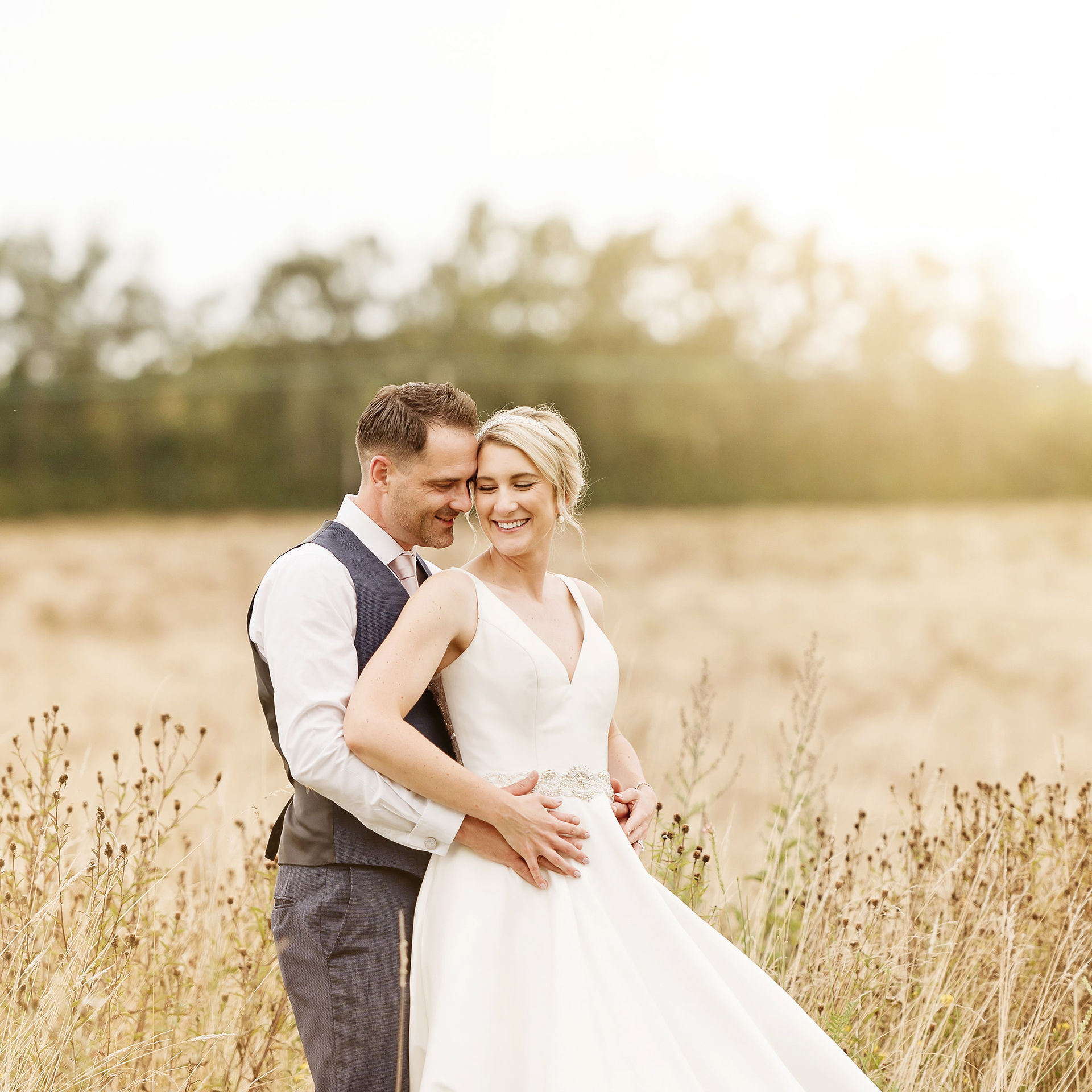 Groom hugging the bride from behind while both smile in a golden field beside Glewstone Court Country House
