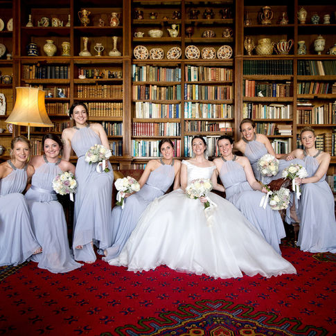 A bride in a white wedding dress sits on a vintage sofa surrounded by her seven bridesmaids in light lavender dresses, all holding bouquets. Captured by Herefordshire Wedding Photographer Russell Lewis at Homme House, they are positioned in front of a large bookcase filled with books and decorative items, with a red patterned rug underneath