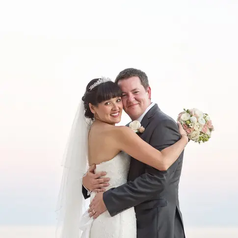 A natural wedding portrait of a bride and groom holding each other beside the sea in Dubrovnik, Croatia, with a gentle sunset glowing in the background on their wedding day