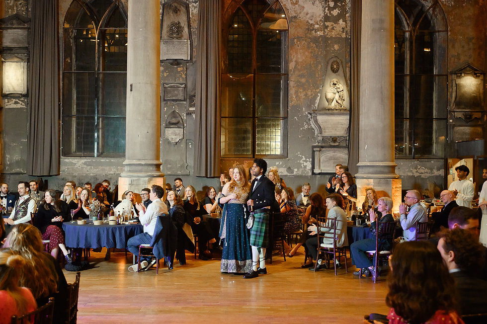 A couple stands in elegant attire at a gathering in a grand hall with large windows and columns, surrounded by seated, applauding guests.