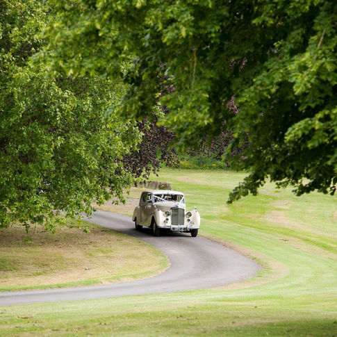 A vintage white car drives along a winding path through a lush, green park with large trees and well-manicured grass at Burton Court. The car is partially obscured by foliage, suggesting a peaceful and serene outdoor setting perfect for capturing by Herefordshire wedding photographer Russell Lewis