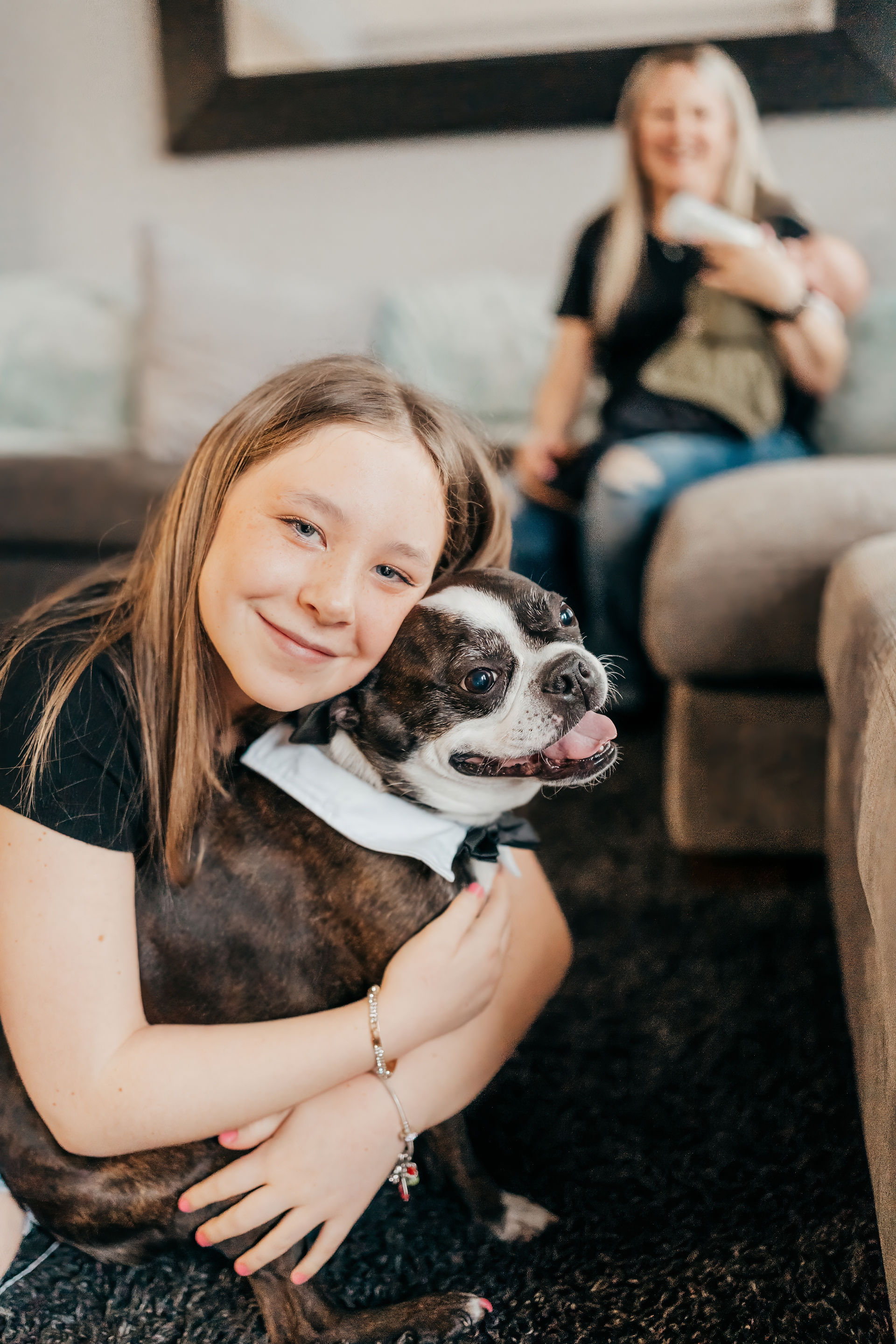 Girl hugging her boston terrier with mom and newborn watching from the background
