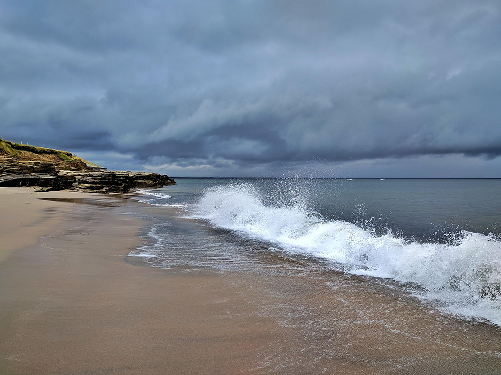 dark grey clouds, stormy weather, splashing waves, dark sea, sandy beach, rocks in the distance. Cresswell, northumberland