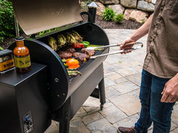 Person grilling steaks, vegetables, and corn on a Traeger wood pellet grill outdoors