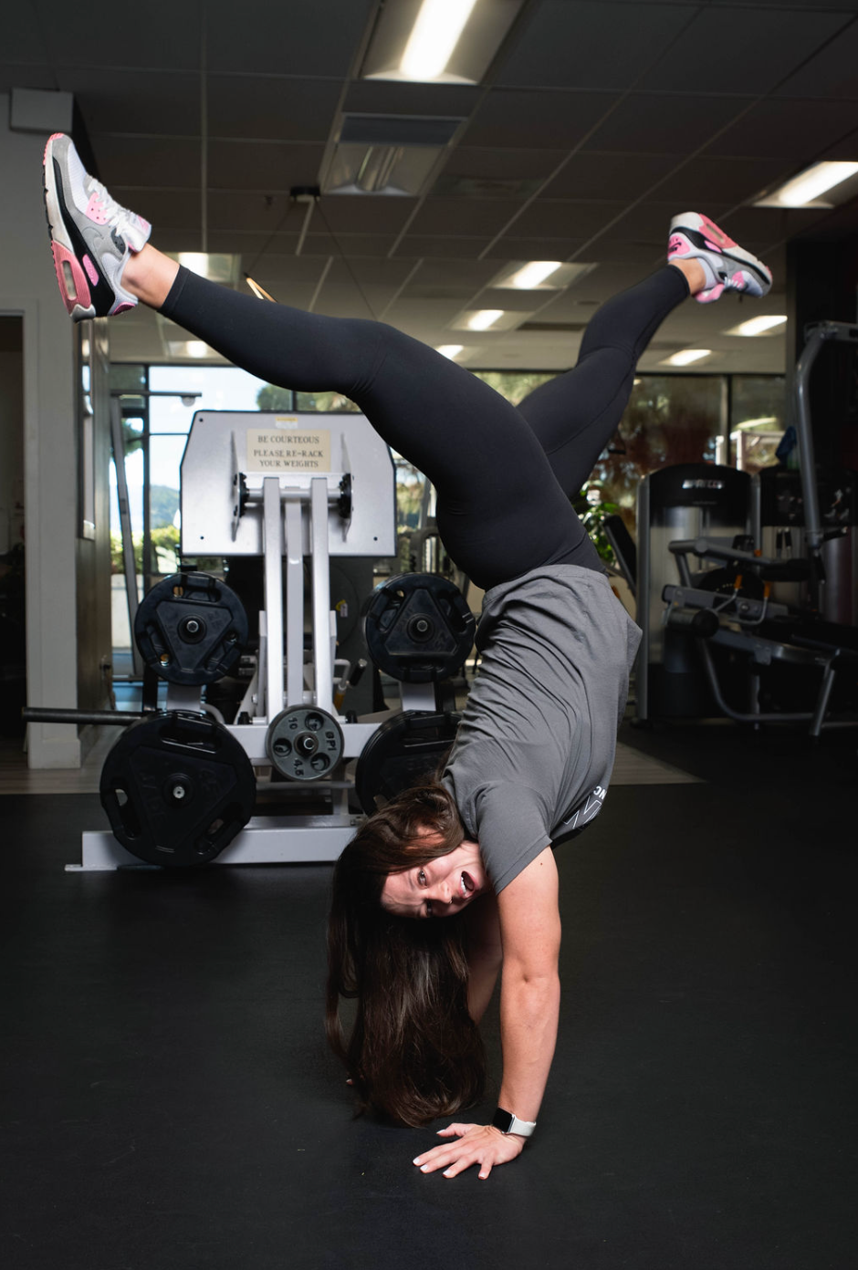 Woman doing a handstand in a gym, smiling. Black leggings, pink and gray sneakers. Gym equipment in the background. Text: "Be courteous."