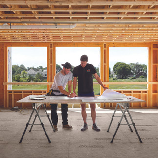 Two men study blueprints on a wooden table in a partially constructed house. Open framework with a view of greenery and houses outside.