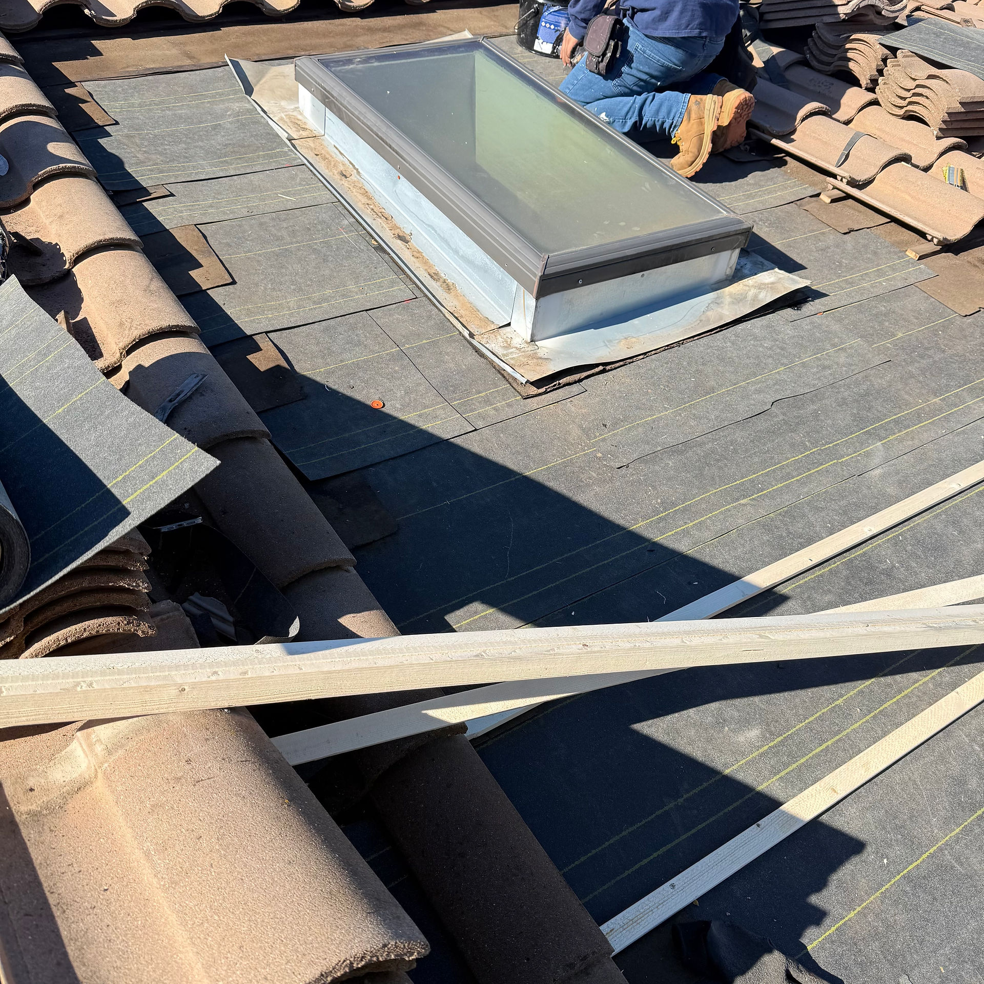 Worker on roof installing skylight surrounded by brown tiles. Sunlit area with shadows, blue jeans and brown boots visible.