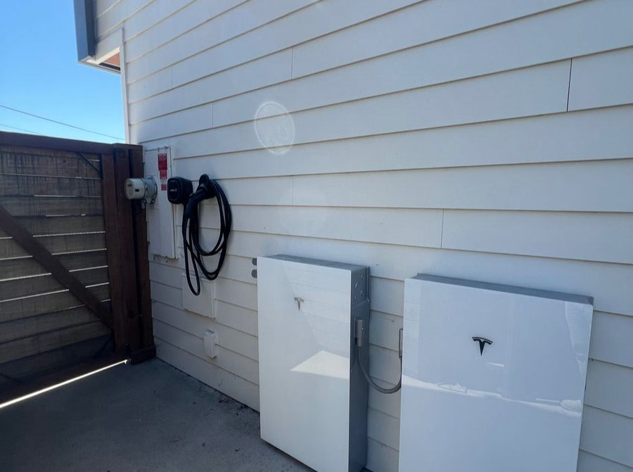White Tesla Powerwalls and an EV charger on a light-colored exterior wall, under a clear blue sky, with sunlight reflecting.