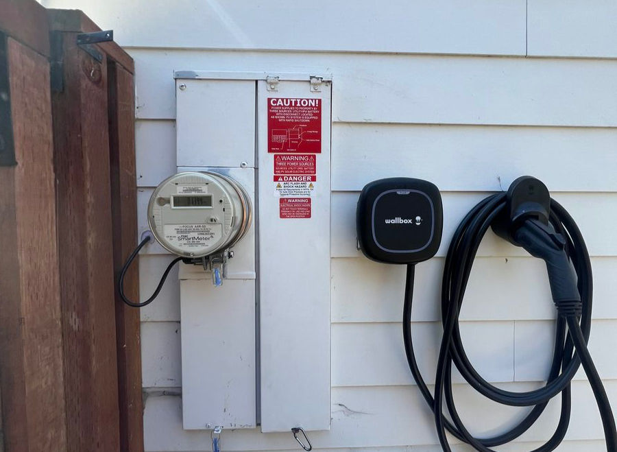 Electric meter and EV charger mounted on a white house wall. Includes a red caution sign. Coiled charging cable hangs from the unit.
