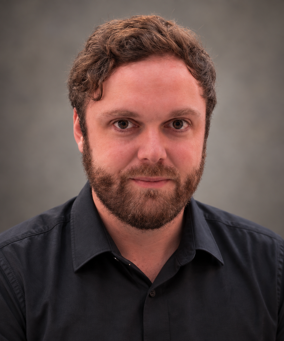 Portrait of a bearded man with curly hair in a dark shirt against a gray background, looking calmly at the camera