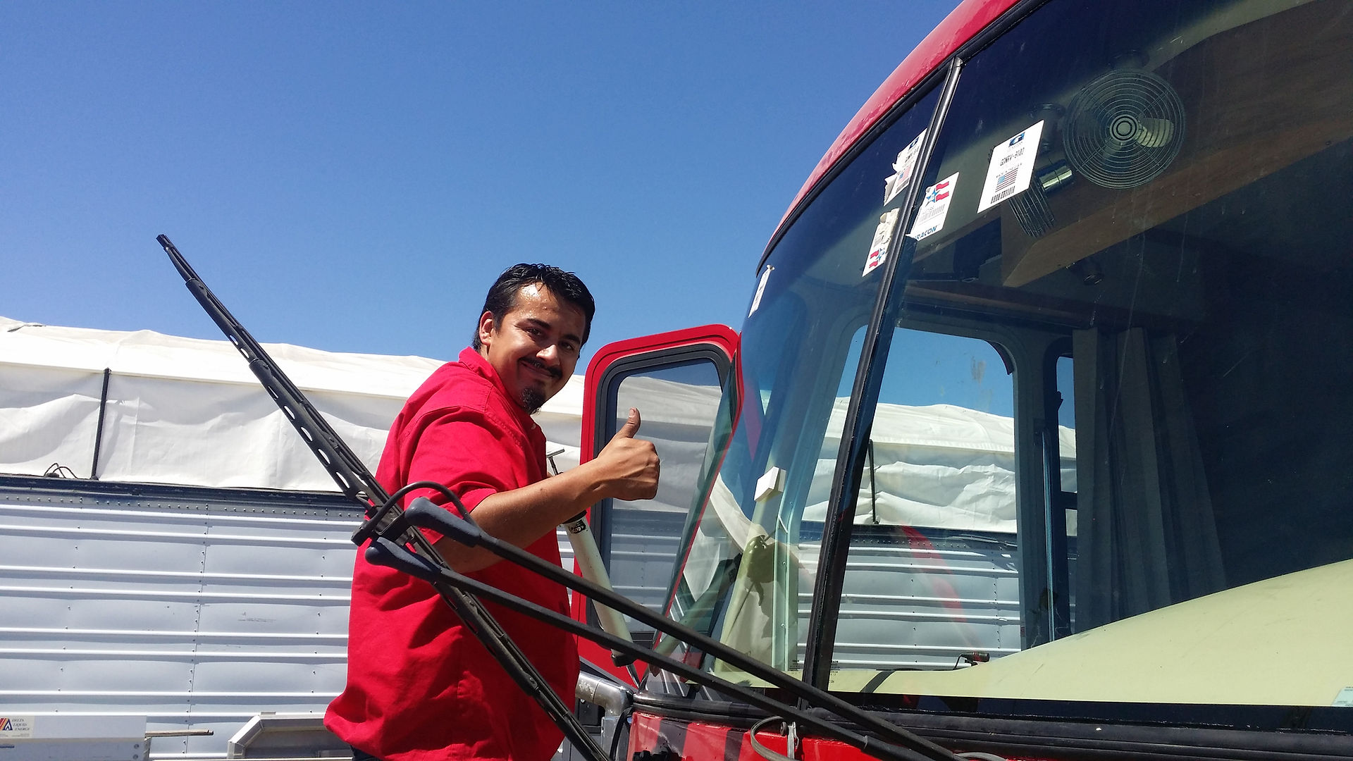 Man in red shirt gives thumbs up, smiling by a red vehicle. Clear blue sky and white tarp in background. Positive mood.