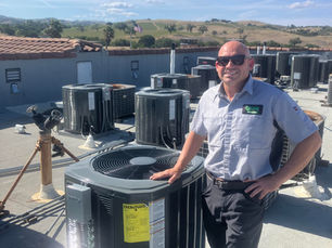 Smiling man in sunglasses stands next to rooftop HVAC units. Clear, sunny day with hills in the background. Wears a uniform with "QUINN" logo.