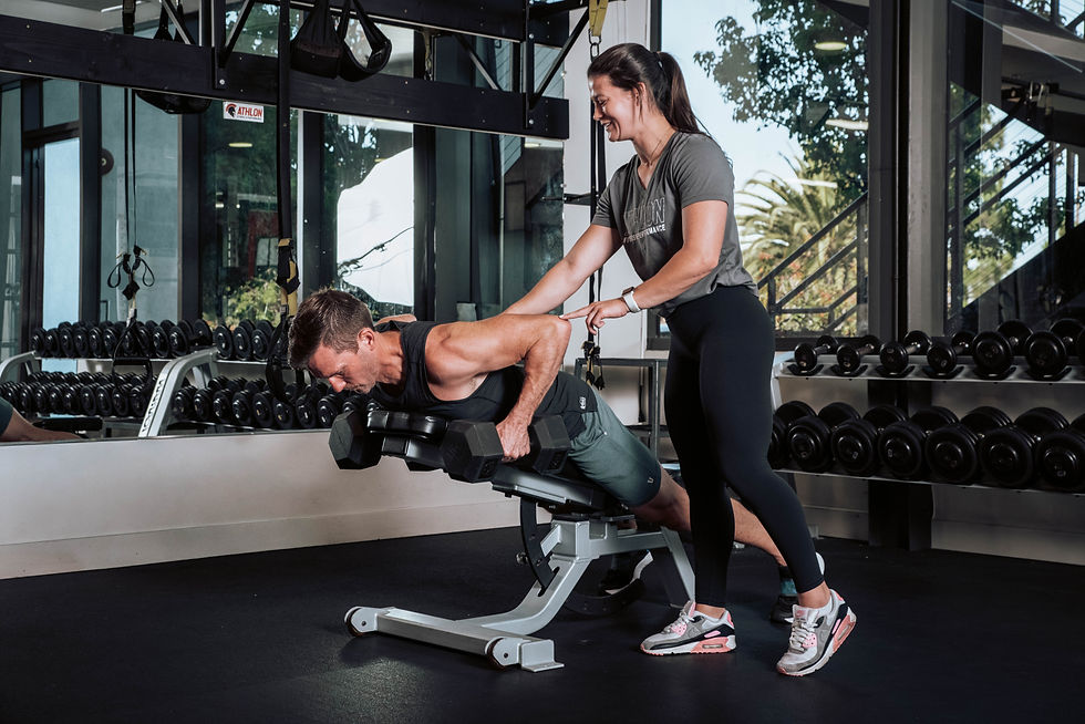 Man lifting weights on an incline bench while a woman assists, smiling. Gym setting with dumbbells and mirrors in the background.