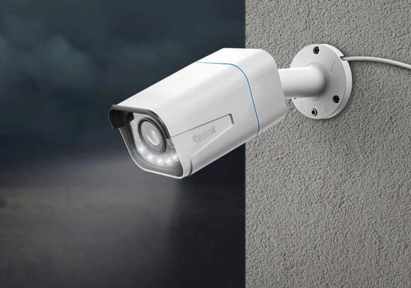 White security camera labeled "Reolink" mounted on a textured wall, facing cloudy night sky. Illuminated LED lights surround the lens.
