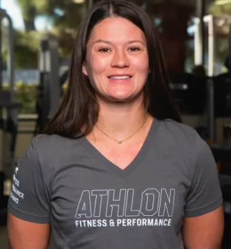 Smiling woman in a gray ATHLON FITNESS & PERFORMANCE T-shirt stands indoors with a blurred gym background.