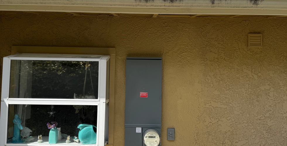 Outdoor deck with metal chairs and table, a covered grill, and beige wall with a window. Overcast sky and power cables above. New panel is installed and the wall is painted to match the house.